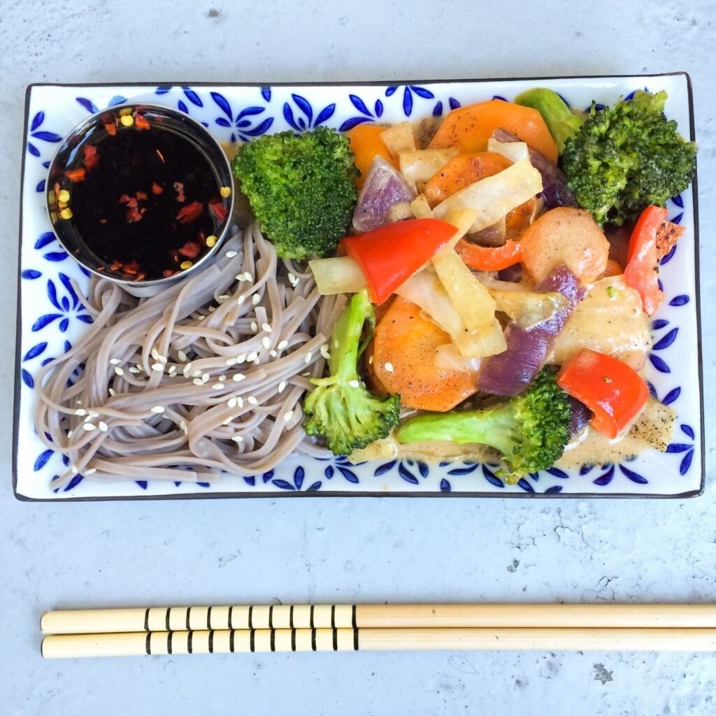 High protein foods for vegetarians featuring a healthy vegetable stir-fry with broccoli, carrots, bell peppers, onions, soba noodles, sesame seeds, and soy dipping sauce.